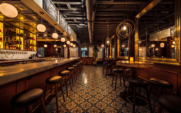 Upscale cocktail bar with marble bar top, globe lighting, mirrored walls, and patterned tile floor.